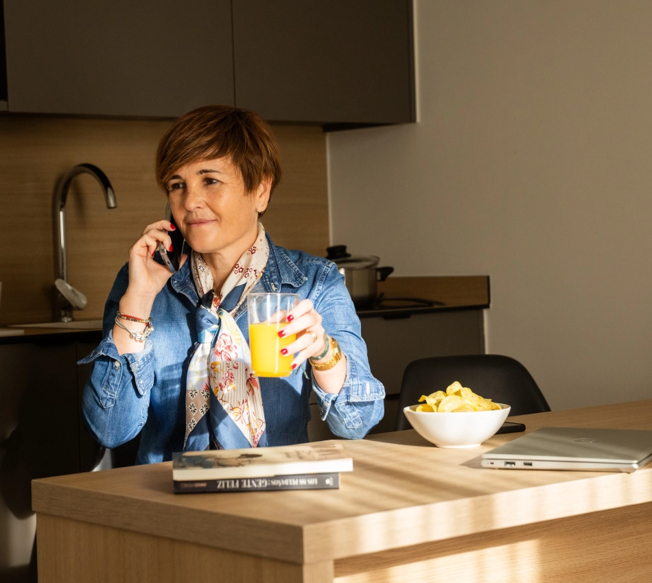 Mujer hablando por tel&eacute;fono y bebiendo jugo en una cocina moderna.