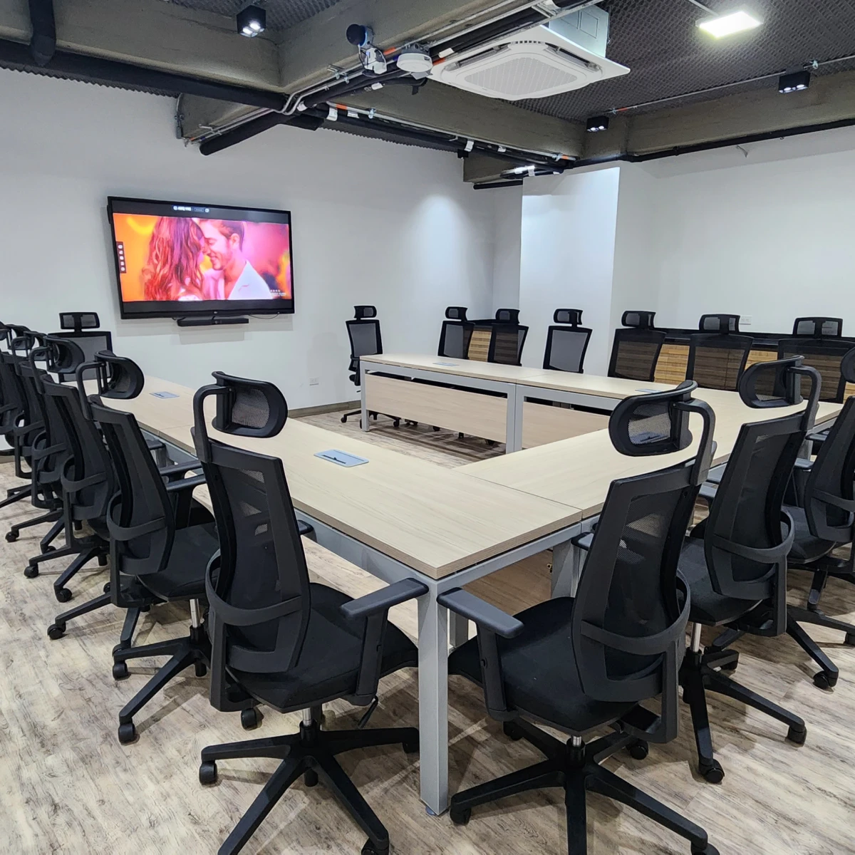 Sala de reuniones con mesas, sillas y pantalla grande encendida.