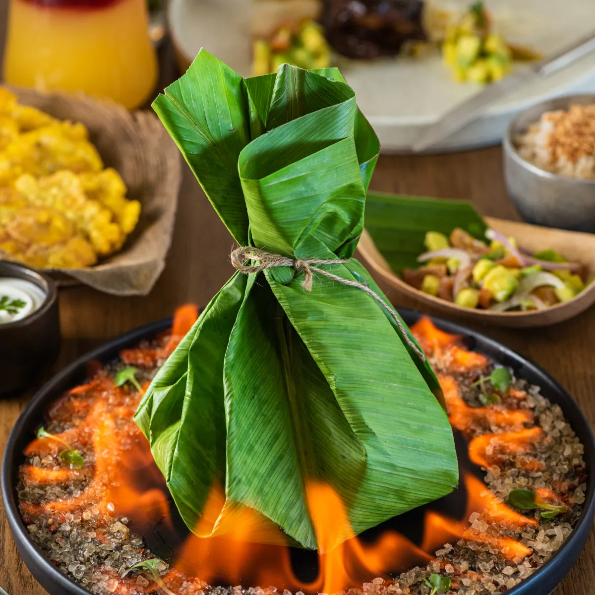 Paquete de hojas sobre un plato con fuego; comida variada alrededor.