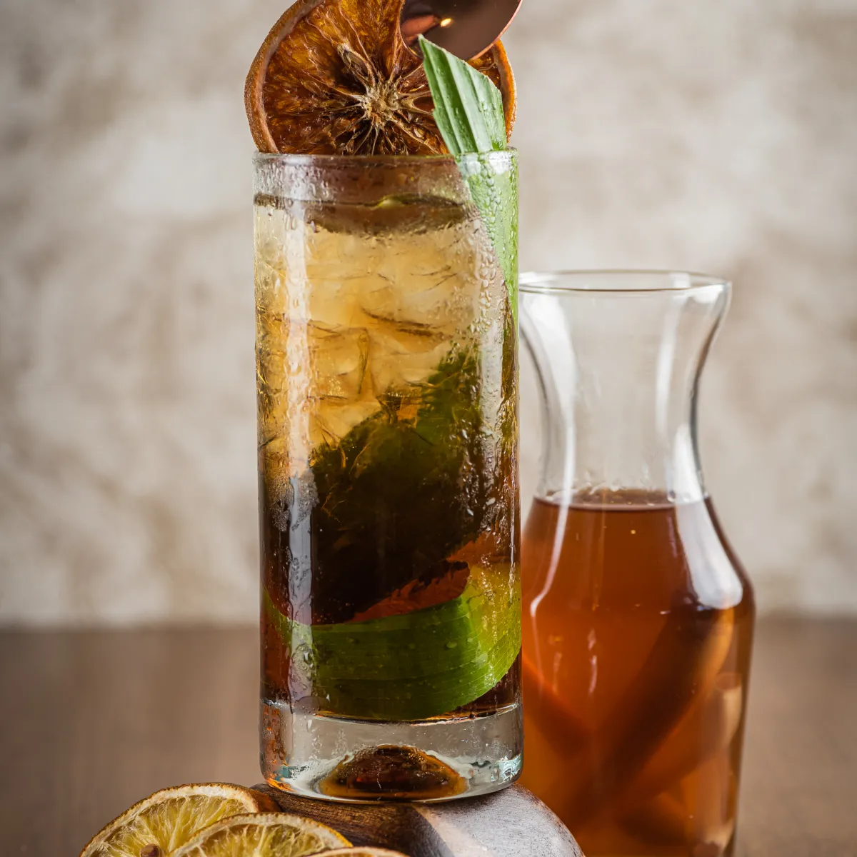 Vaso de cóctel con rodaja de cítrico, junto a jarra con bebida ámbar.