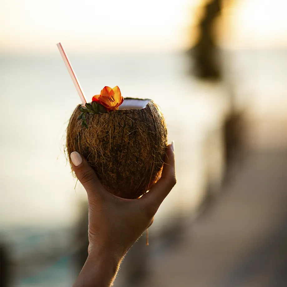 Mano sosteniendo un coco con bebida y flor, fondo borroso de playa al atardecer.