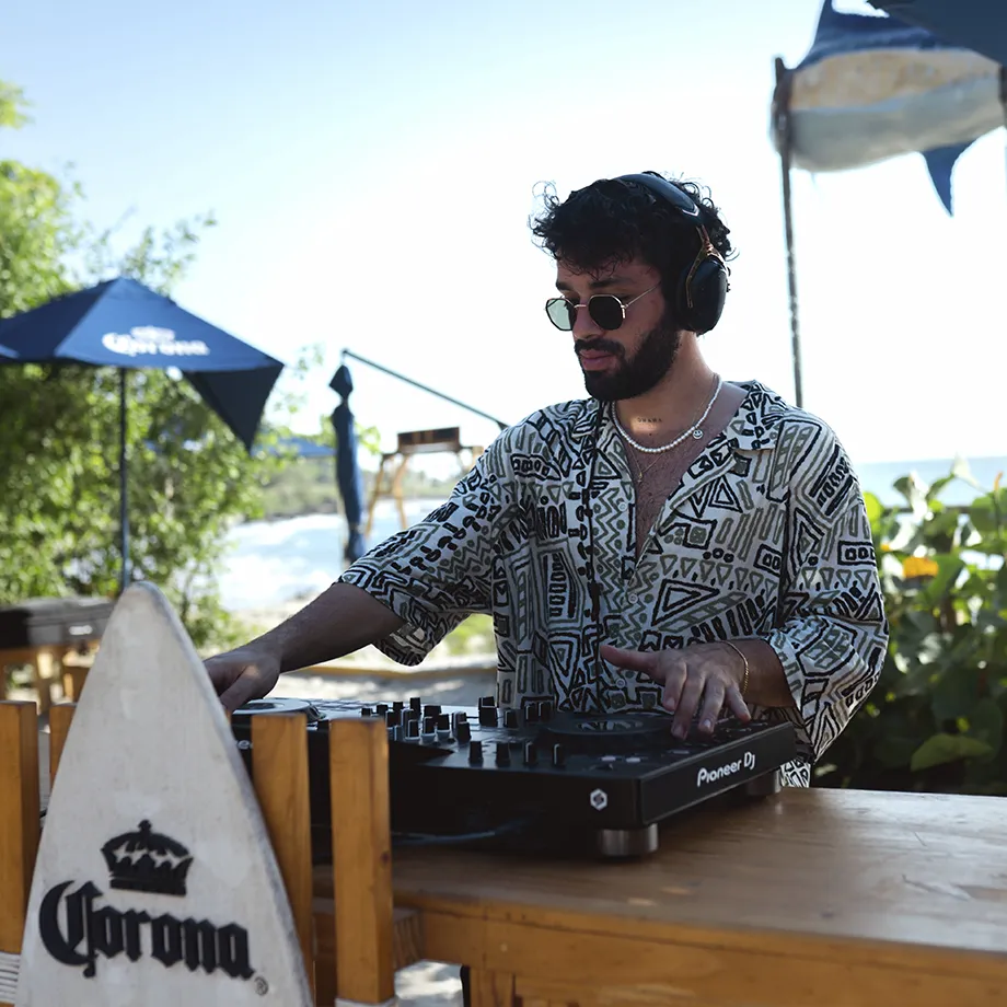 DJ mezclando música en la playa, con vegetación de fondo.