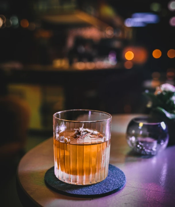 Vaso de whisky con hielo en mesa de bar iluminada.