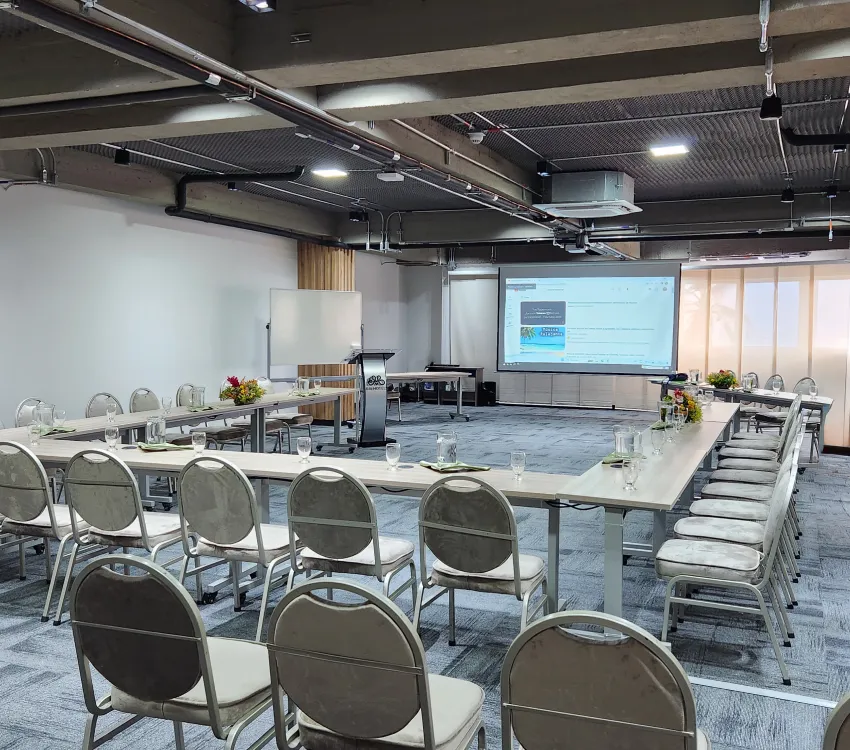 Sala de conferencias vac&iacute;a con disposici&oacute;n de mesas en forma de U y proyector.