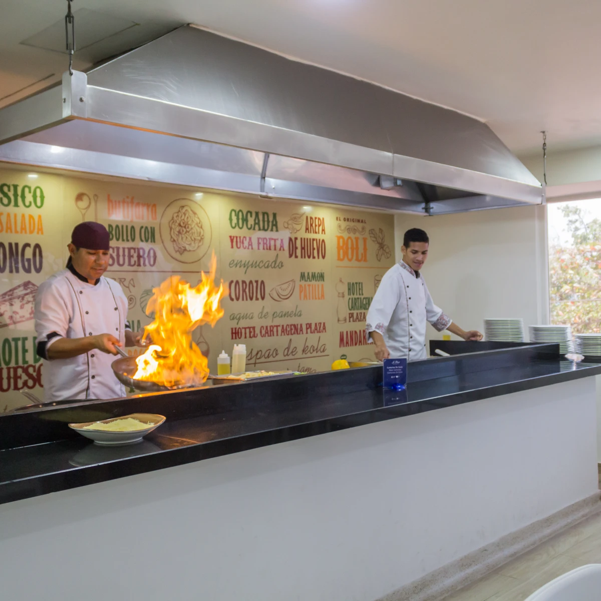 Chef cocinando con llama en cocina abierta, otro chef observando.
