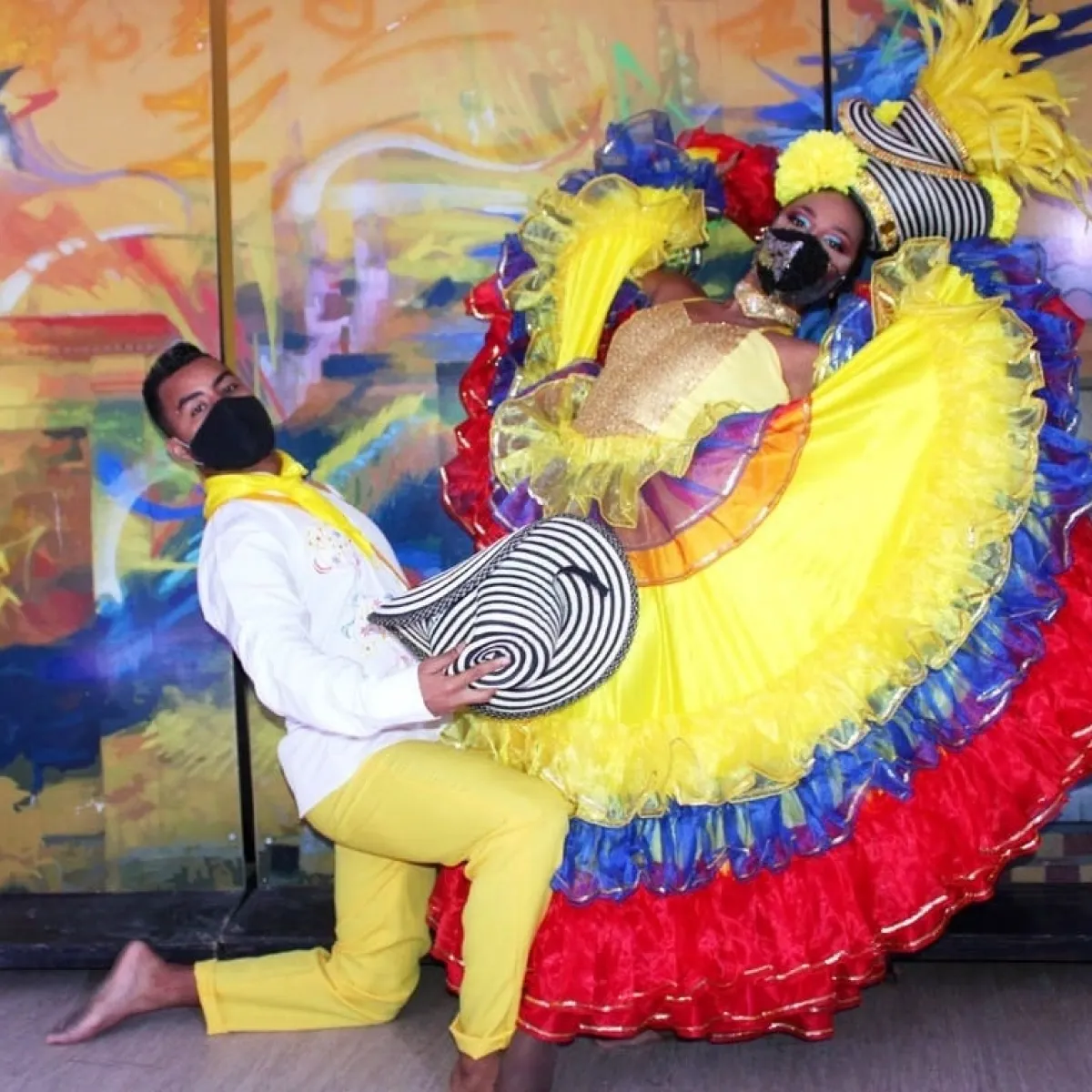 Pareja con trajes coloridos bailando, ambos usan sombreros y mascarillas.