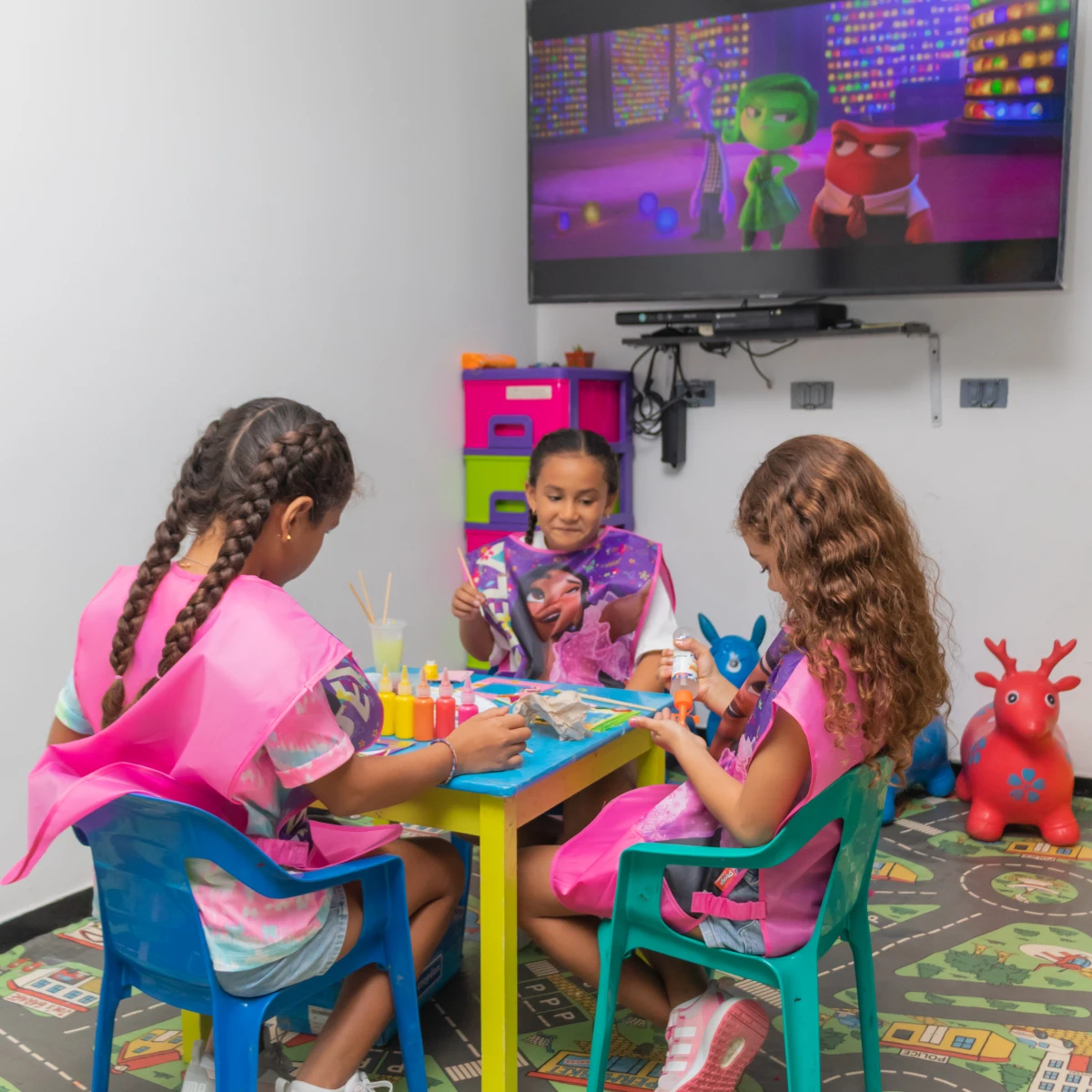 Niñas pintando en una mesa con dibujos animados en la pantalla.