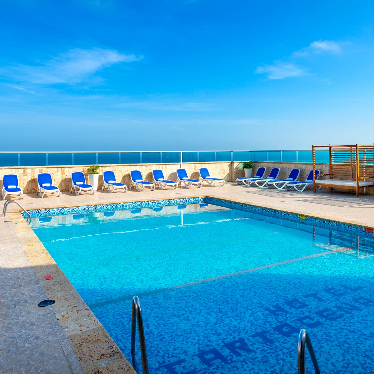 Piscina al aire libre con sillas azules y vista al mar bajo cielo despejado.
