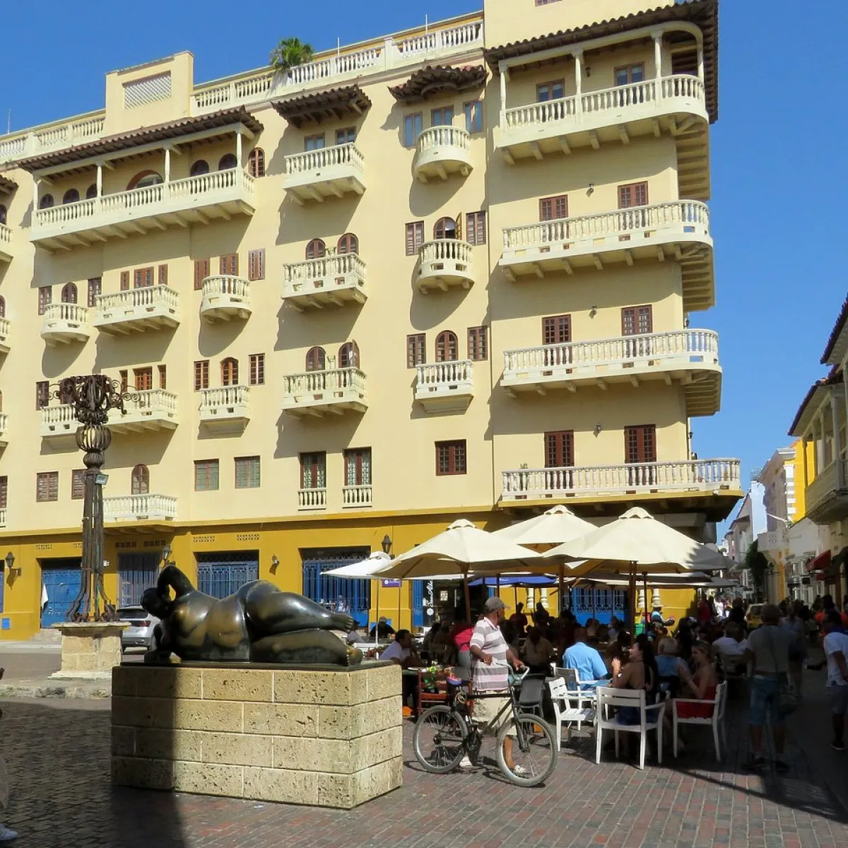 Edificio amarillo con balcones y escultura en calle llena de personas y bicicletas.