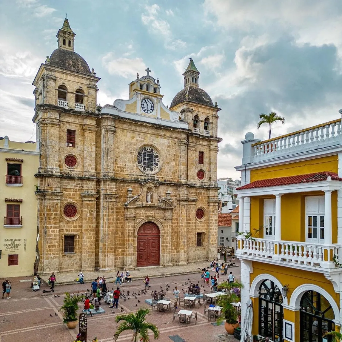 Iglesia colonial y plaza concurrida en Cartagena, frente a edificio amarillo.