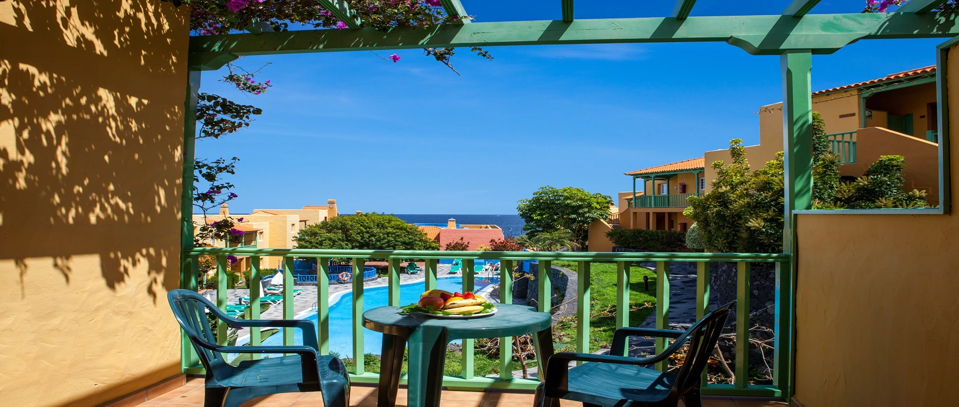 Balc&oacute;n con vista al jard&iacute;n, piscina y mar, con mesa, sillas y frutas.