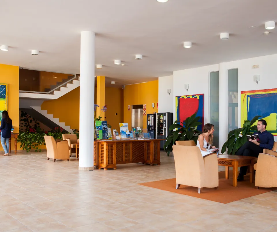 Vest&iacute;bulo colorido con personas conversando en sillones. Hay una escalera al fondo.