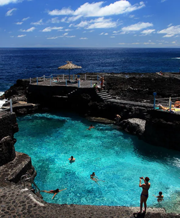 Piscina natural rocosa junto al oc&eacute;ano con gente nadando bajo cielo azul.
