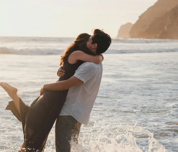 Pareja abraz&aacute;ndose en la orilla del mar al atardecer, con olas rompiendo a su alrededor.