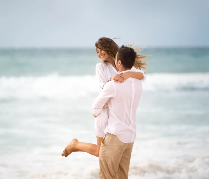 Pareja sonriente en la playa; &eacute;l la levanta en un abrazo mientras el viento mueve su cabello.