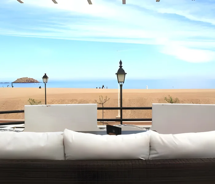 Vista de una playa desde una terraza con sillones blancos y l&aacute;mparas de pie negras.