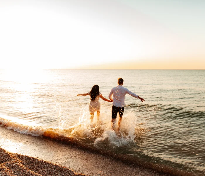 Pareja corre hacia el mar al atardecer, salpicando agua en la orilla.