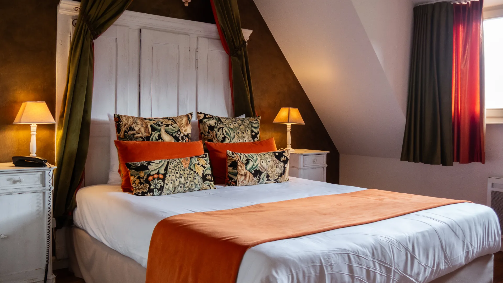 Chambre chaleureuse avec lit double, coussins orange et deux lampes de chevet allum&eacute;es.