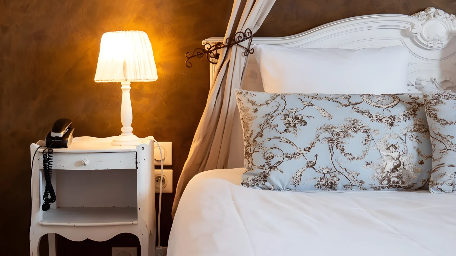 Lampe et t&eacute;l&eacute;phone sur table de chevet &agrave; c&ocirc;t&eacute; d'un lit avec coussin d&eacute;cor&eacute;.