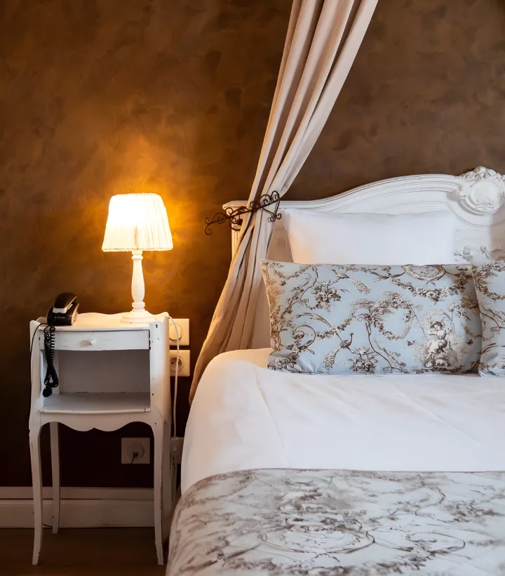 Lit avec oreillers fleuris, table de nuit blanche et lampe allum&eacute;e.