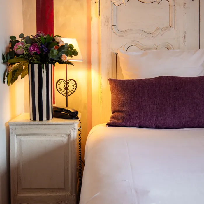 Lit avec coussin violet, table de nuit avec fleurs et lampe allum&eacute;e.