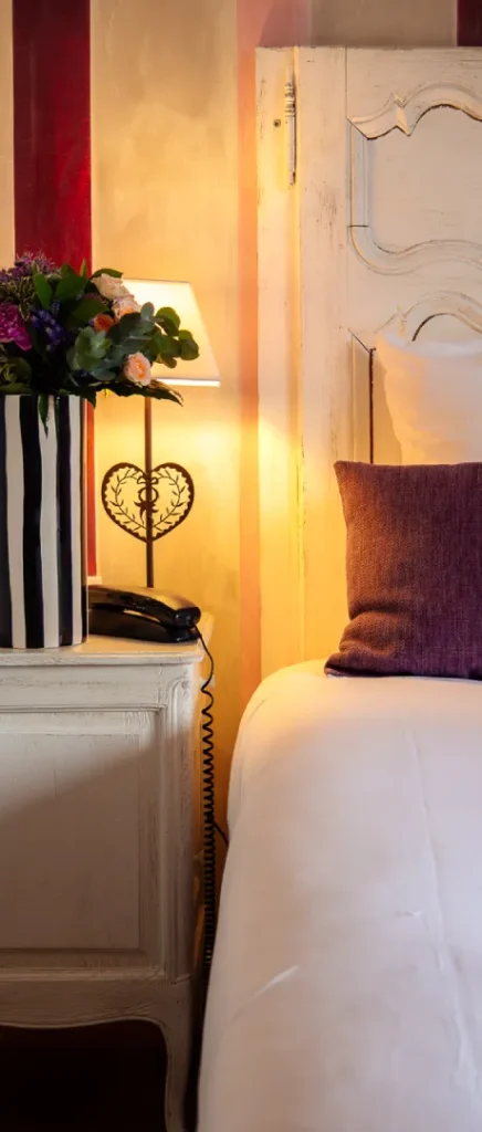 Chevet avec lampe, fleurs et oreiller violet dans une chambre chaleureuse.