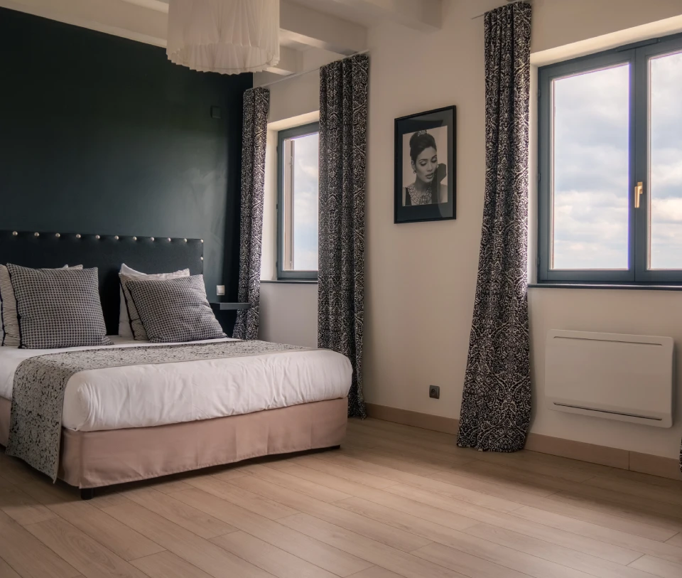 Chambre moderne avec lit double, grandes fen&ecirc;tres et photo encadr&eacute;e au mur.