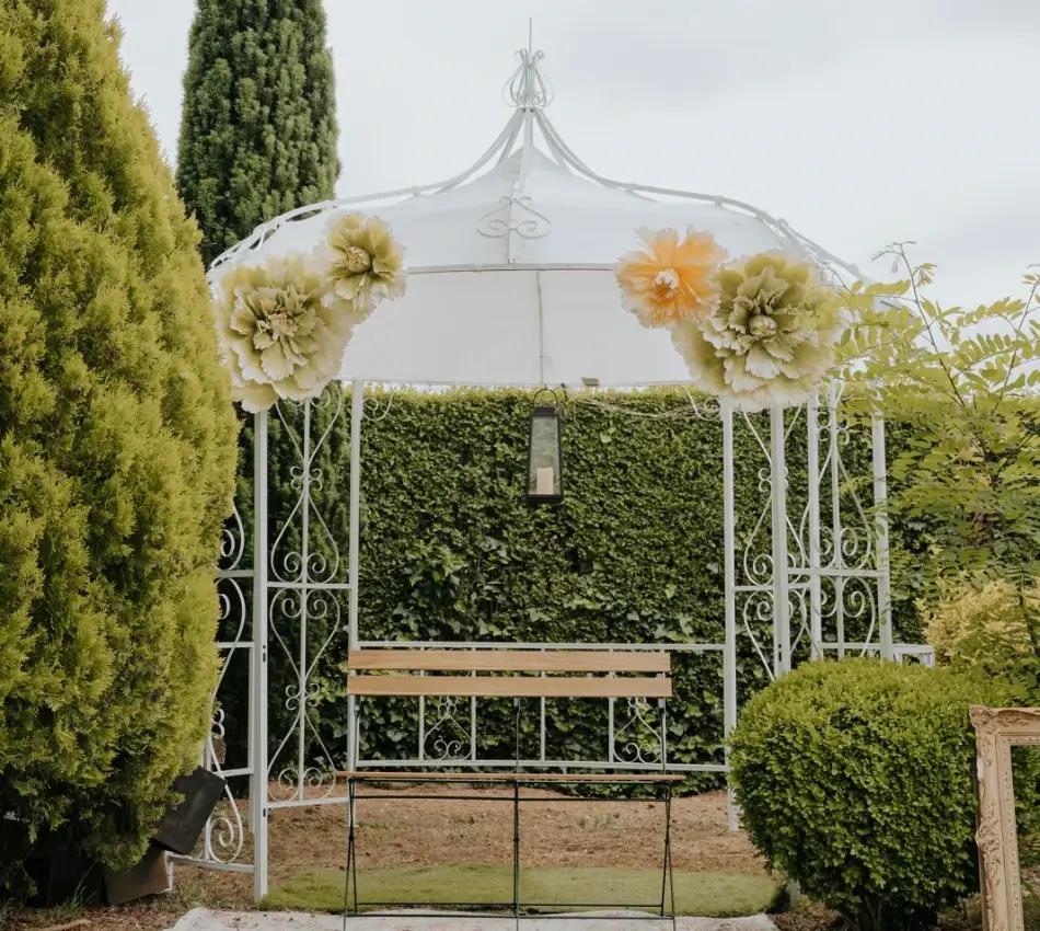 P&eacute;rgola blanca con flores amarillas y verde, seto y banco en jard&iacute;n.