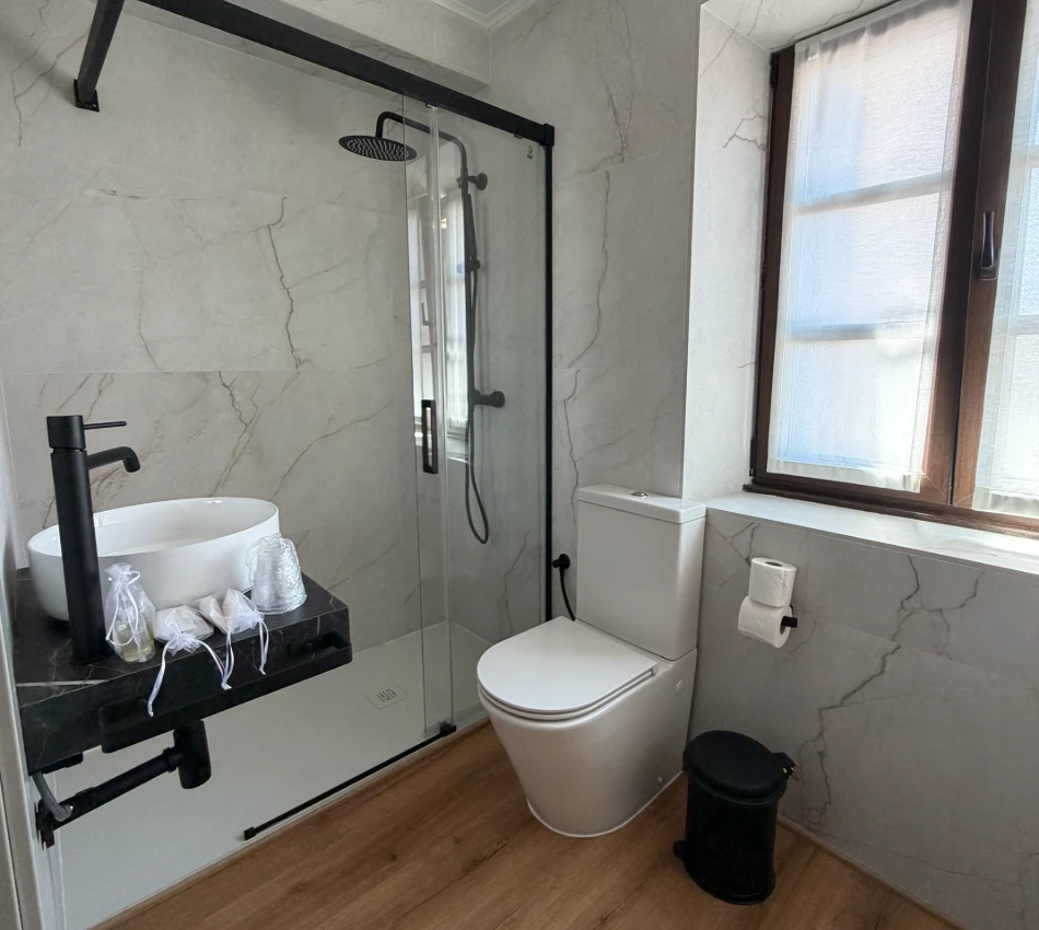 Ba&ntilde;o moderno con ducha de vidrio, inodoro blanco y ventana con cortina.