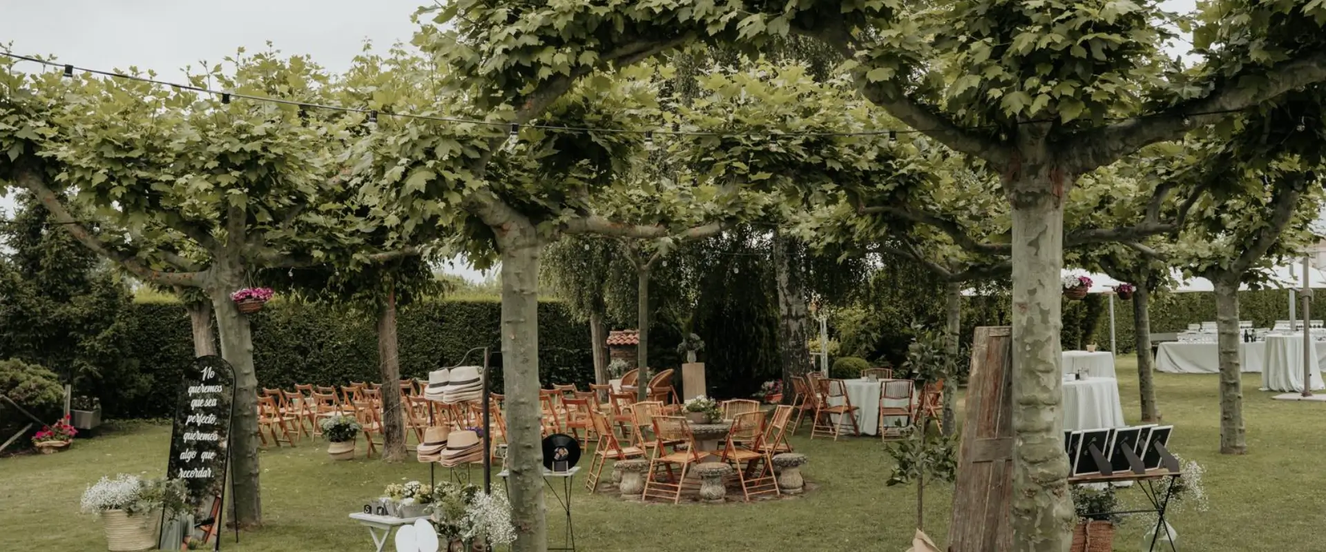 Jard&iacute;n con sillas de madera y mesas, bajo &aacute;rboles y guirnaldas para evento al aire libre