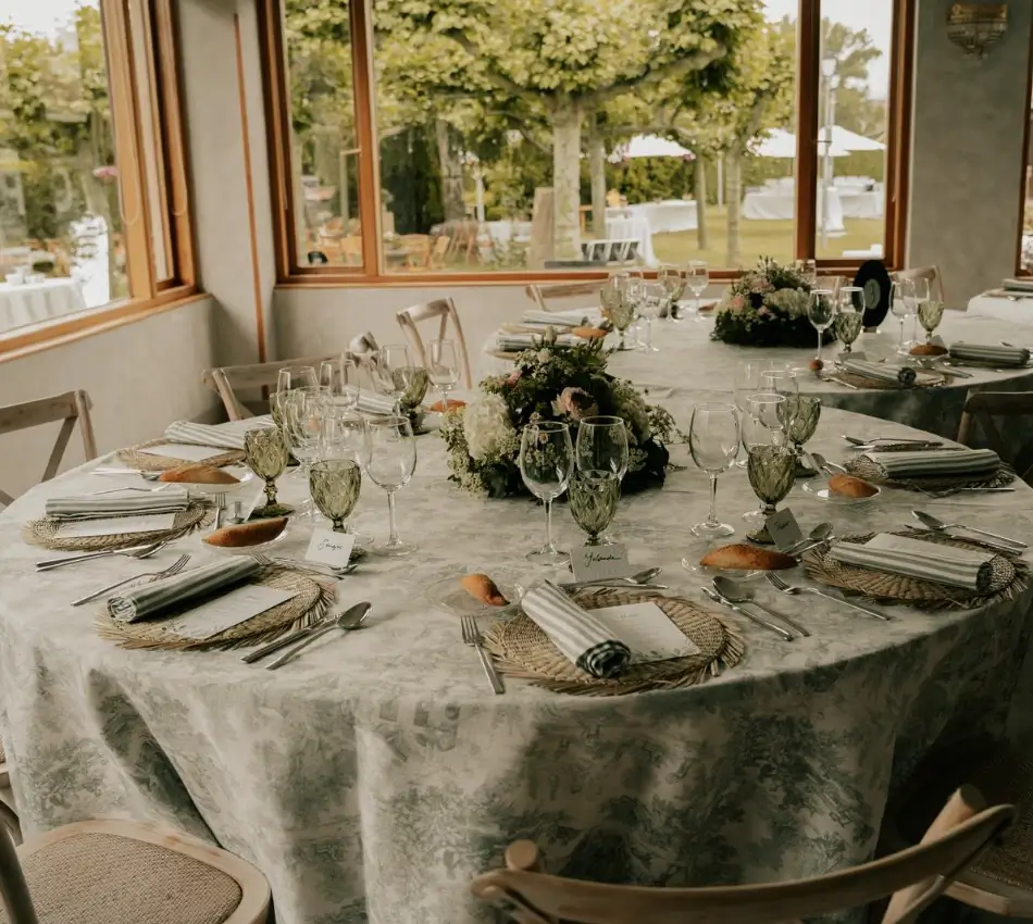Mesa de banquete con copas y arreglos florales en sal&oacute;n con ventanales.