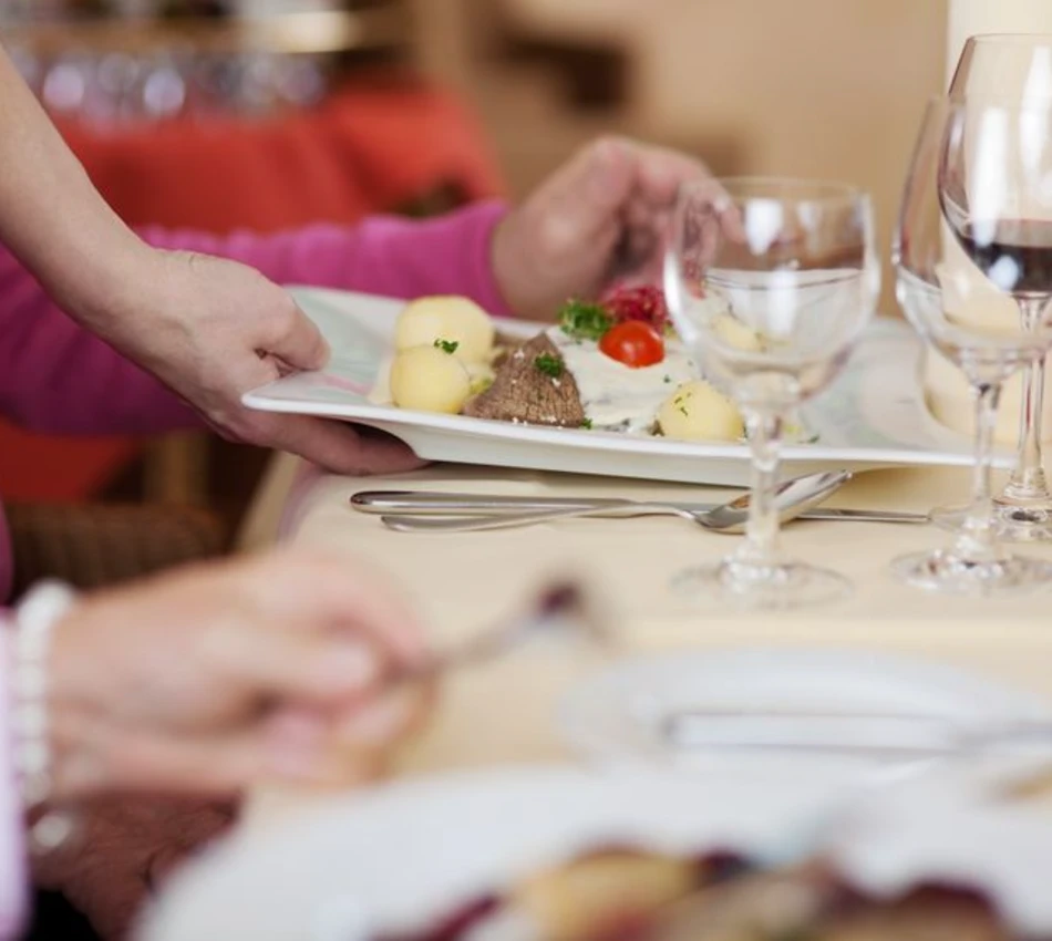 Persona sirviendo plato con papas, carne y tomate en mesa con cubiertos y vino