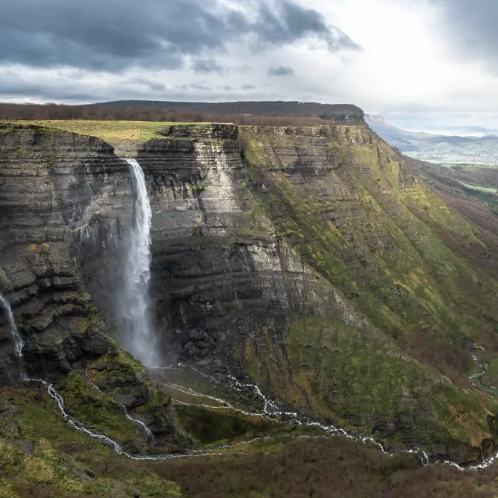 Cascada en un acantilado rocoso con paisaje verde y cielo nublado.