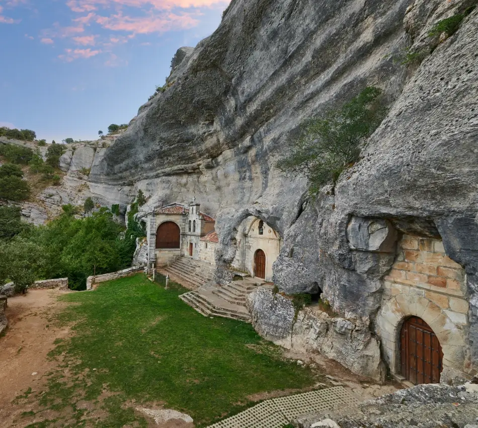 Ermita en cuevas bajo un acantilado rocoso con escaleras y arcos.