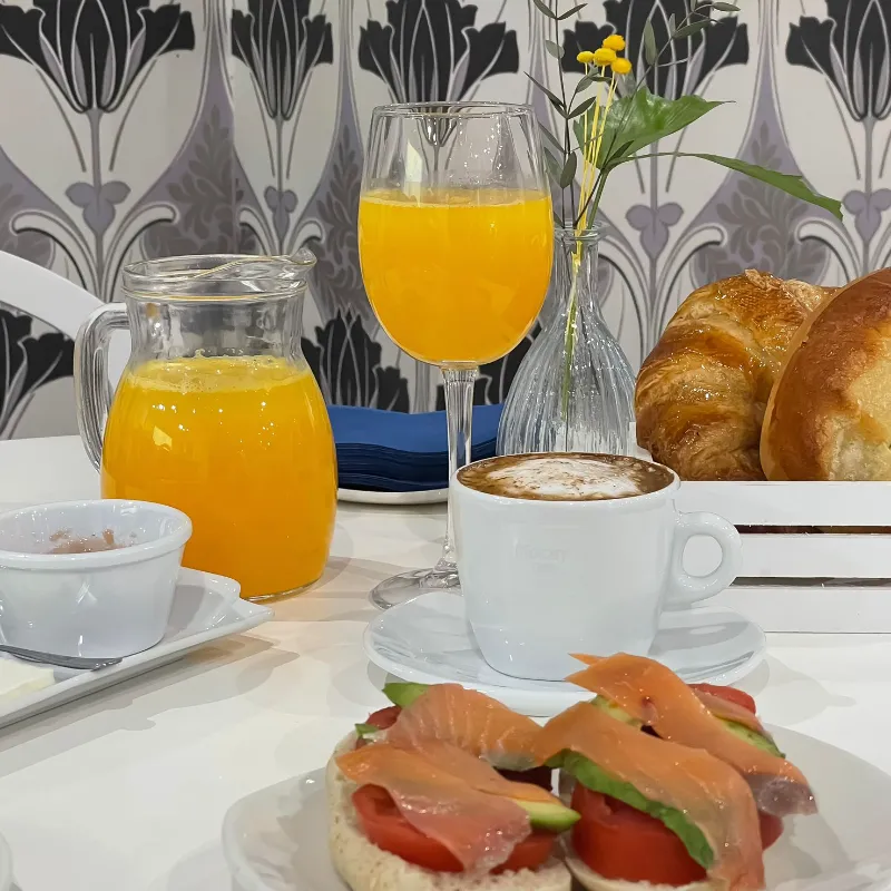 Desayuno con jugo de naranja, capuchino, bagels con salm&oacute;n, croissants y flores en la mesa.