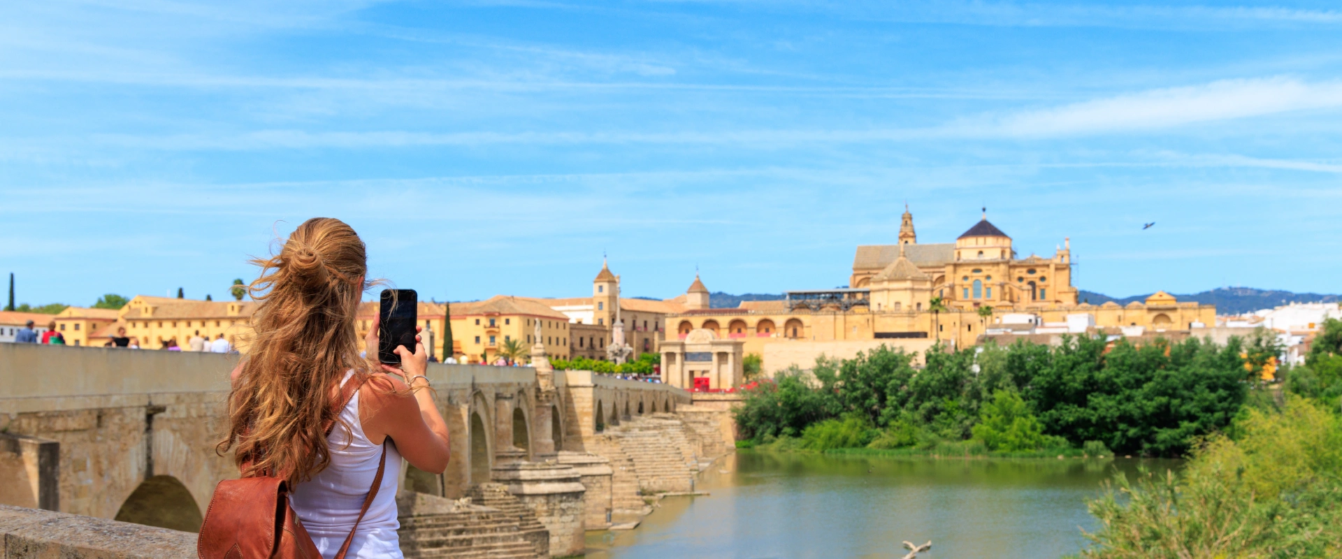 Mujer tomando foto de un puente y un edificio hist&oacute;rico bajo un cielo azul claro.
