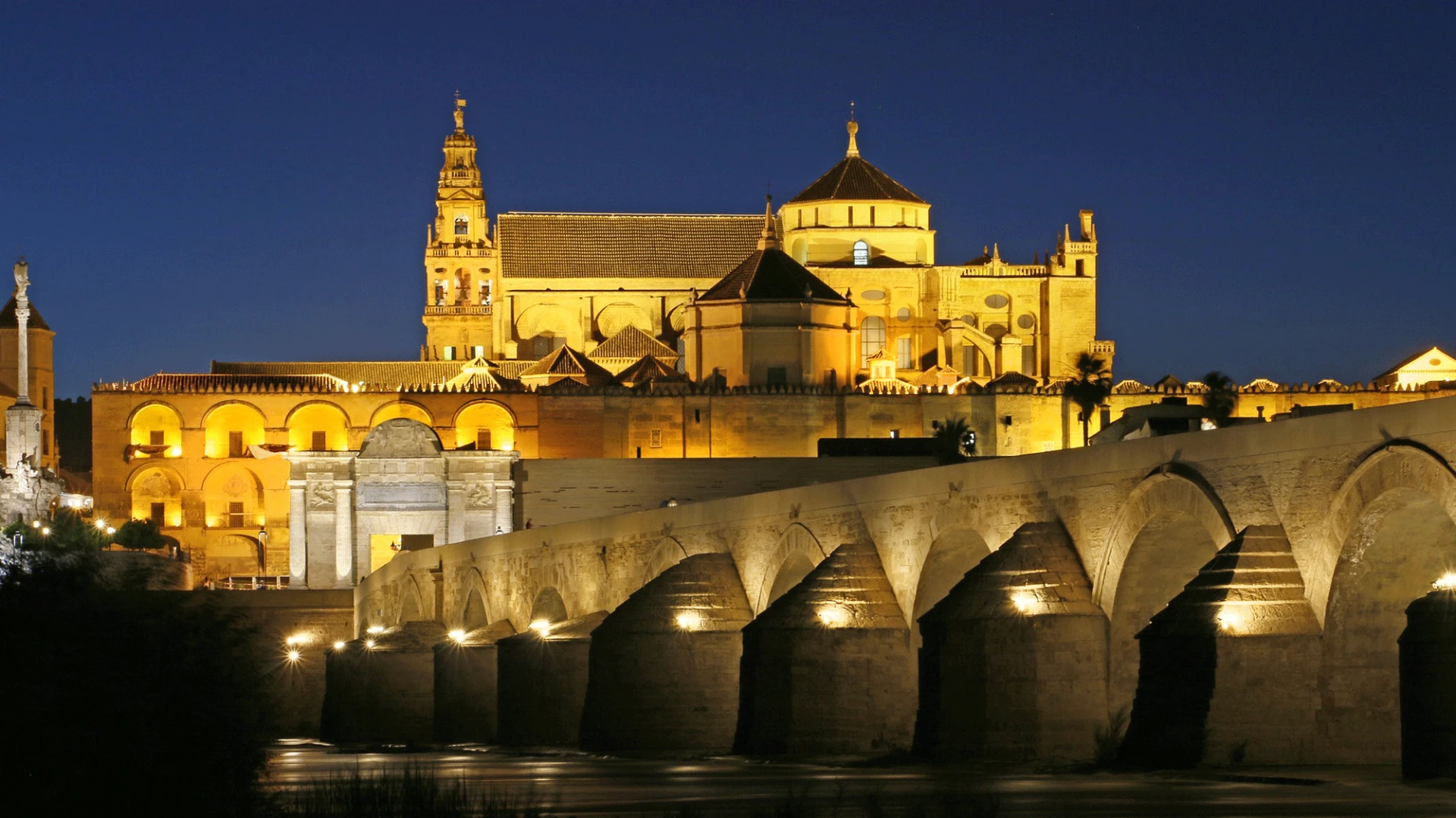 Edificio hist&oacute;rico iluminado con puente en primer plano durante la noche.