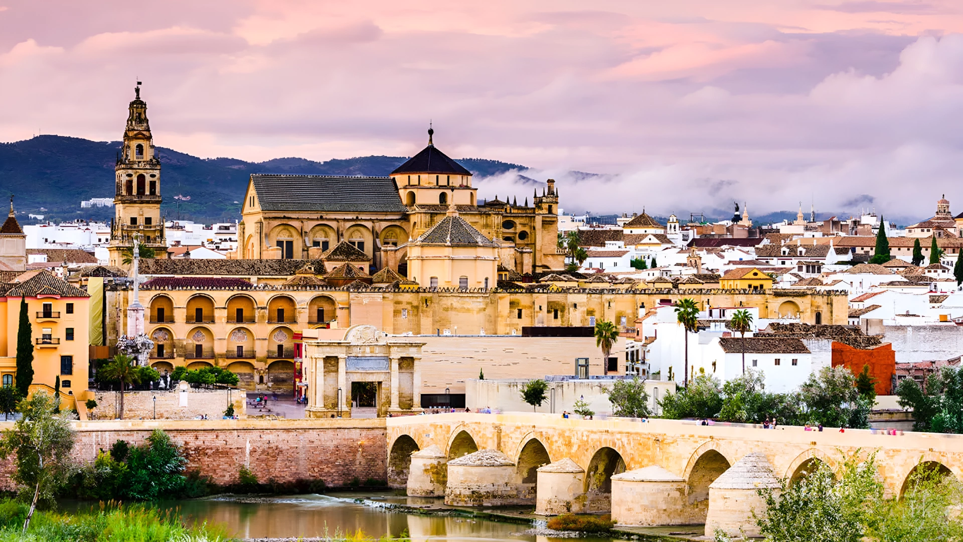 Ciudad hist&oacute;rica con puente y catedral al atardecer, monta&ntilde;as al fondo.