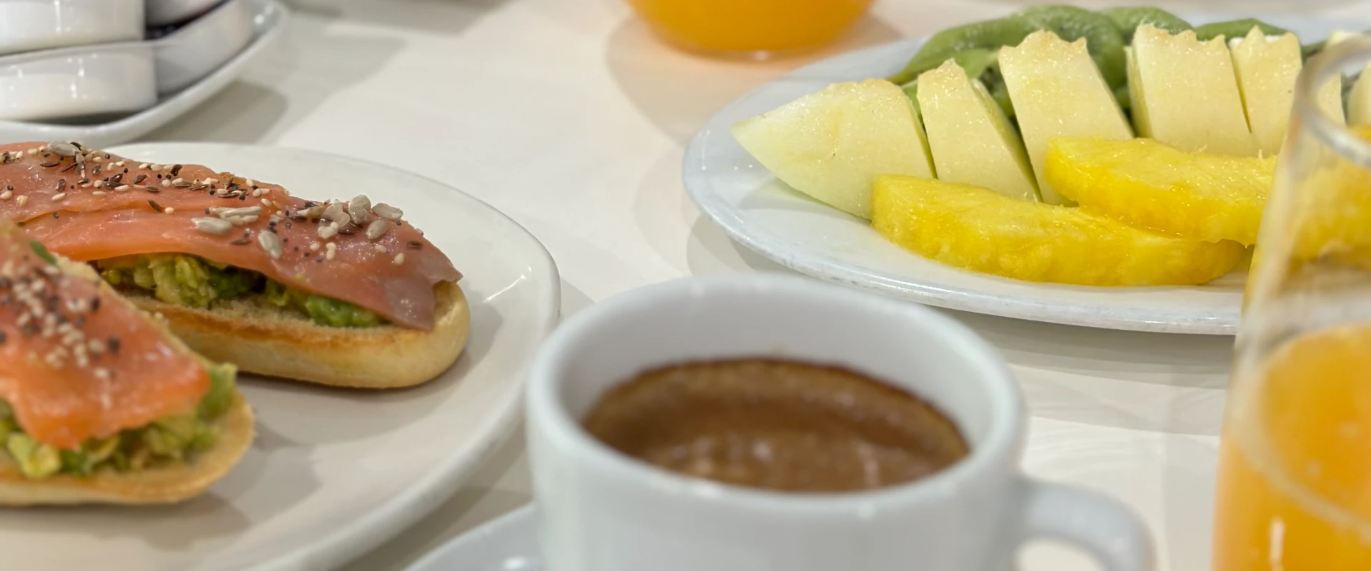 Desayuno con tostada de aguacate y salm&oacute;n, rodajas de fruta y caf&eacute; en una mesa blanca.