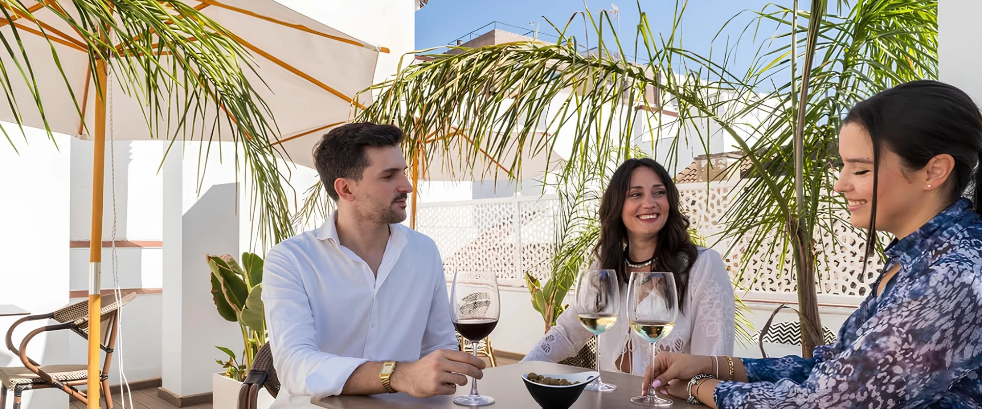 Personas conversando y bebiendo vino en una terraza soleada con plantas y sombrillas.