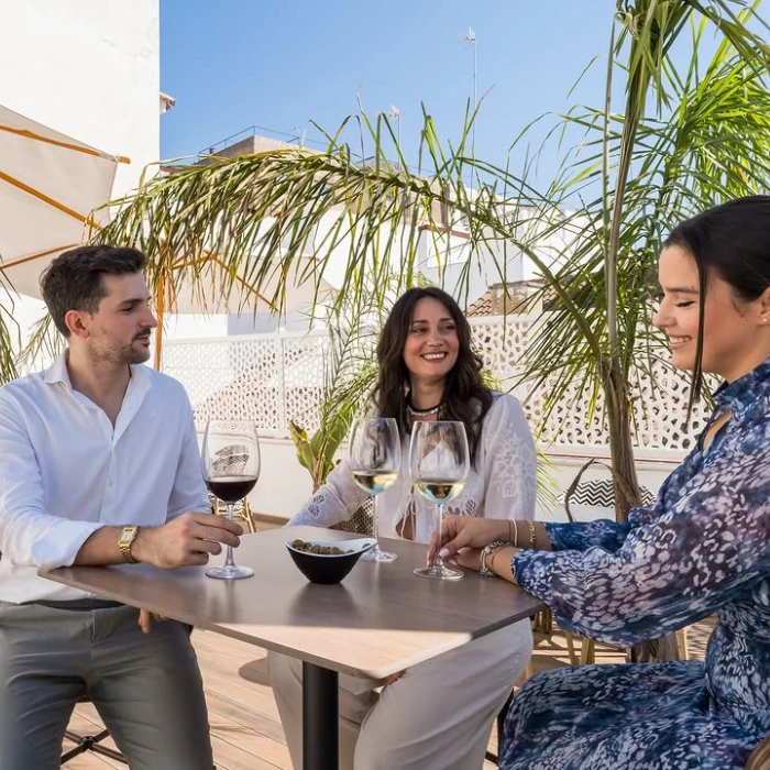 Tres personas beben vino en una terraza soleada con palmeras y charlan felices.