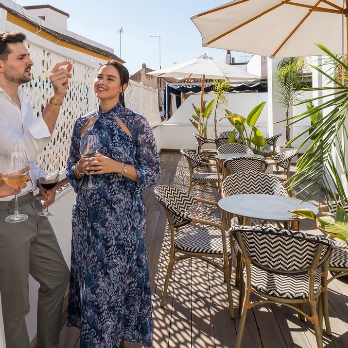Dos personas beben vino en una terraza soleada con sillas, mesas y sombrillas alrededor.