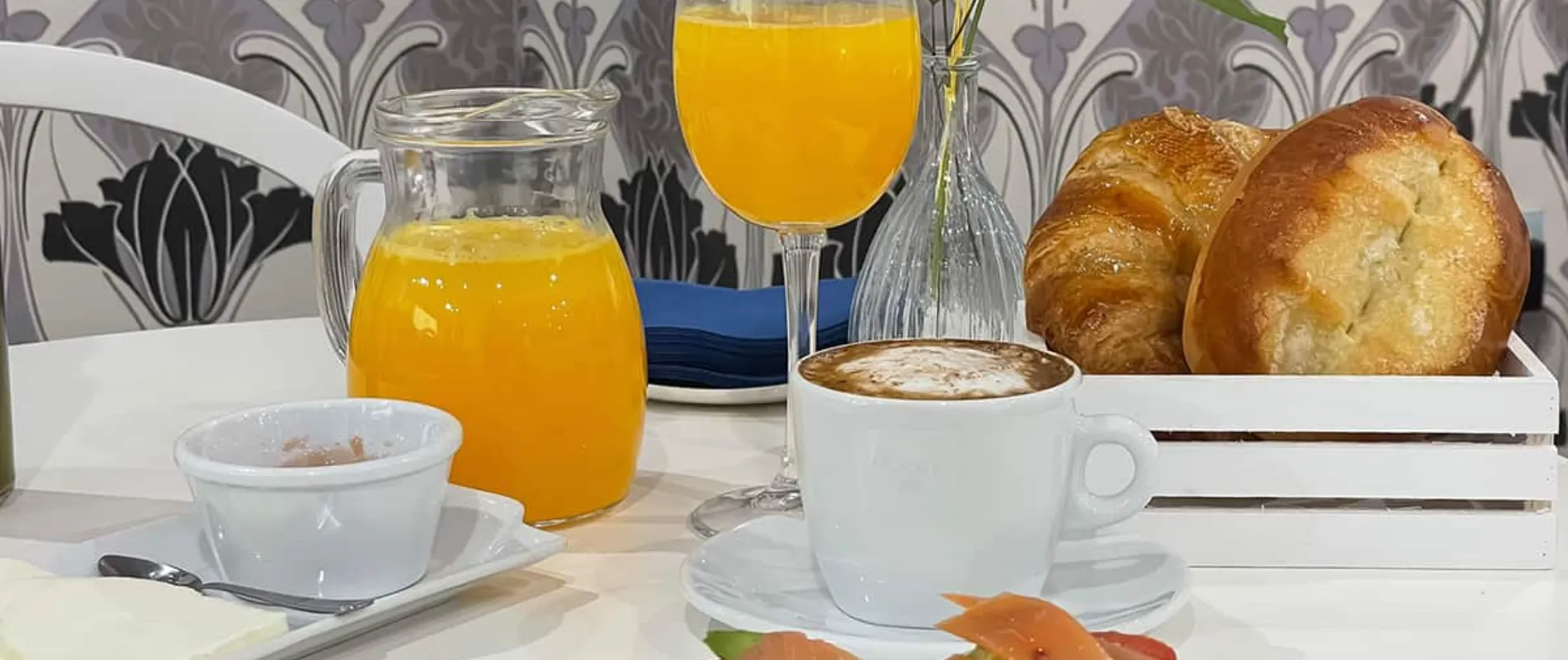 Desayuno con caf&eacute;, jugo de naranja, bollos y un plato con fruta, en un entorno elegante.