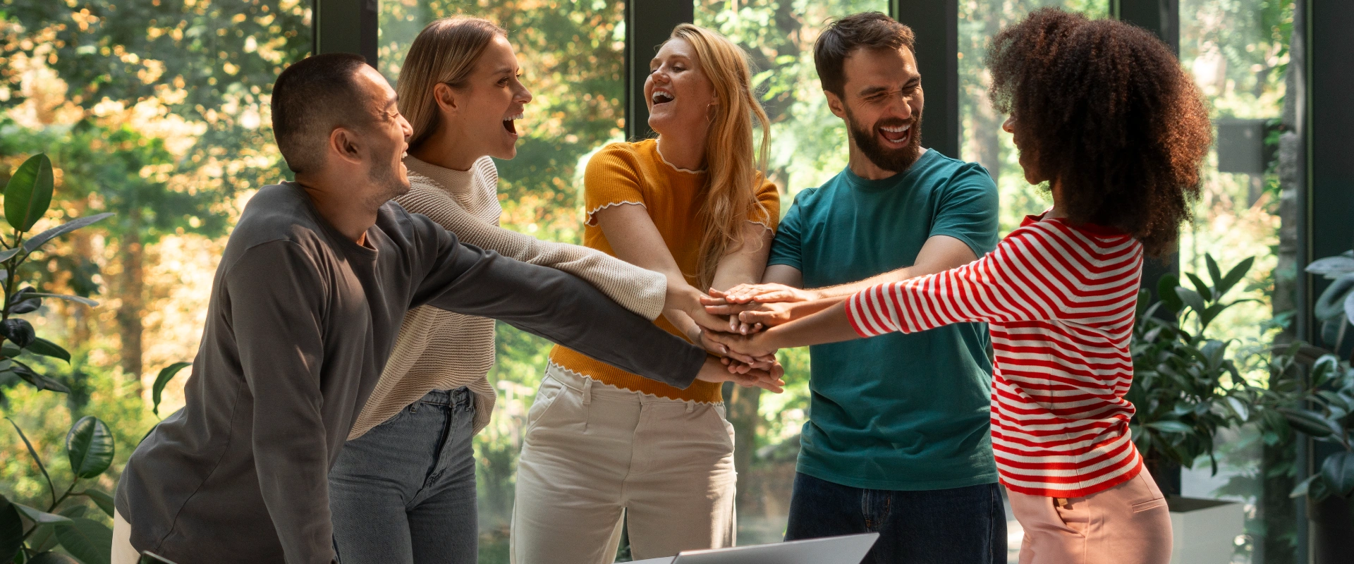 Grupo de personas unidas con las manos en un gesto de trabajo en equipo, sonriendo en un interior verde.