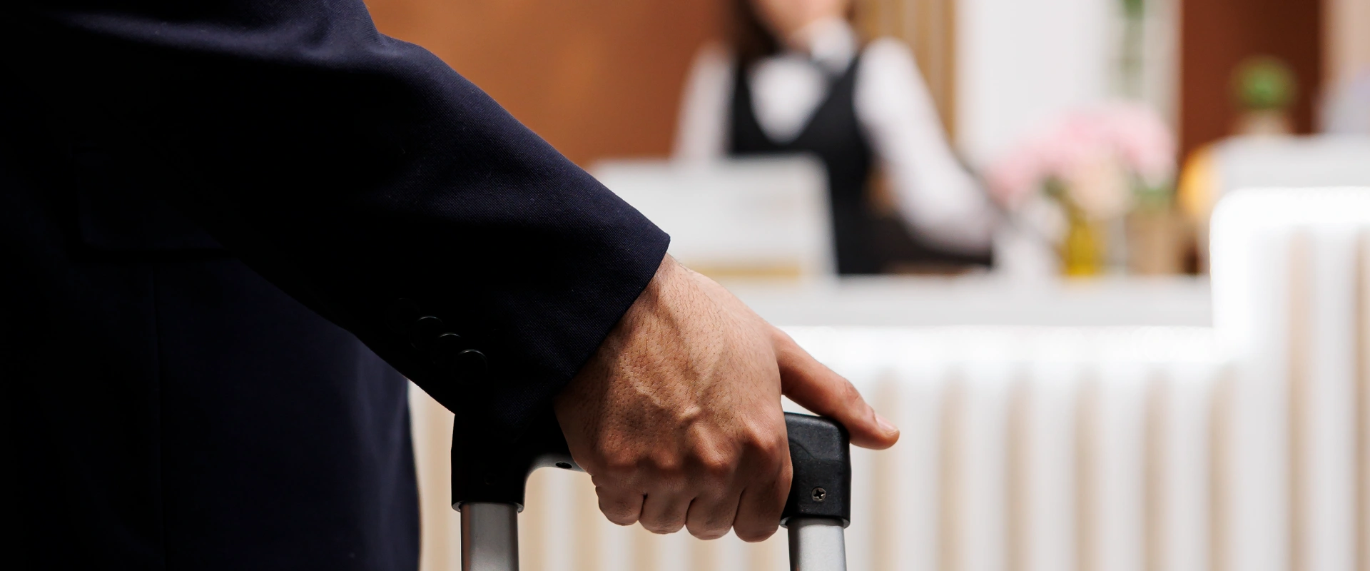 Mano de una persona sujetando una maleta en el lobby de un hotel, recepcionista al fondo.