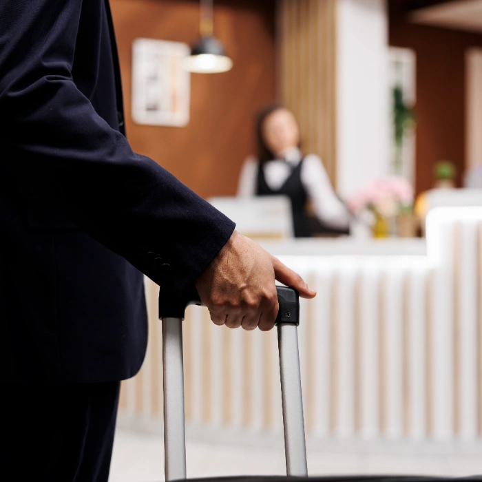 Persona con maleta en recepci&oacute;n de hotel mientras una recepcionista est&aacute; al fondo.