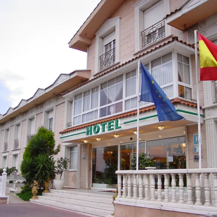 Fachada de hotel con banderas de la UE y Espa&ntilde;a, entrada con columnas blancas y plantas.