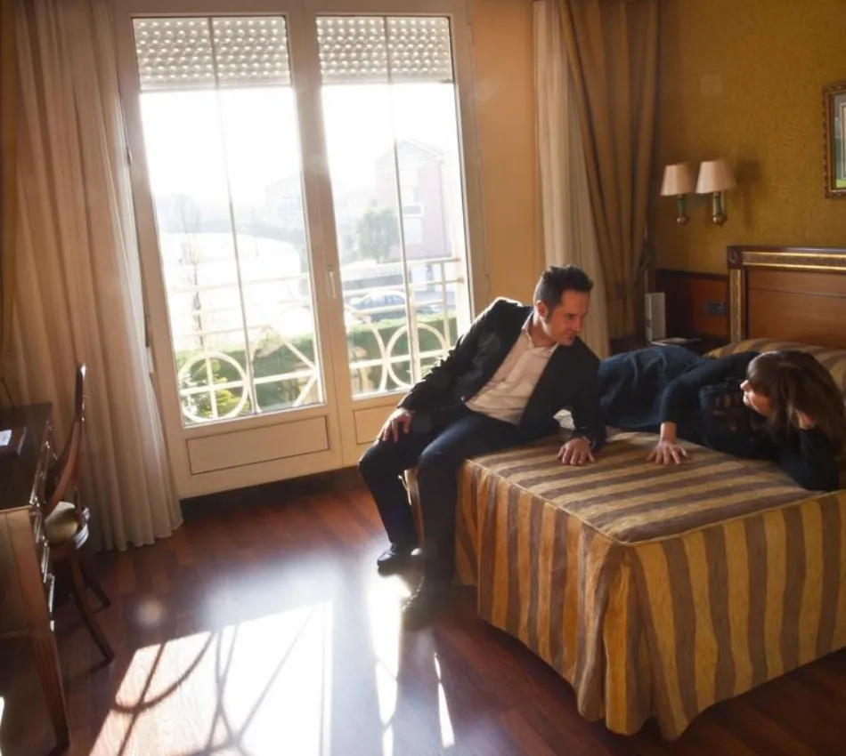 Hombre y mujer conversan en una habitaci&oacute;n iluminada, junto a una ventana grande y soleada.