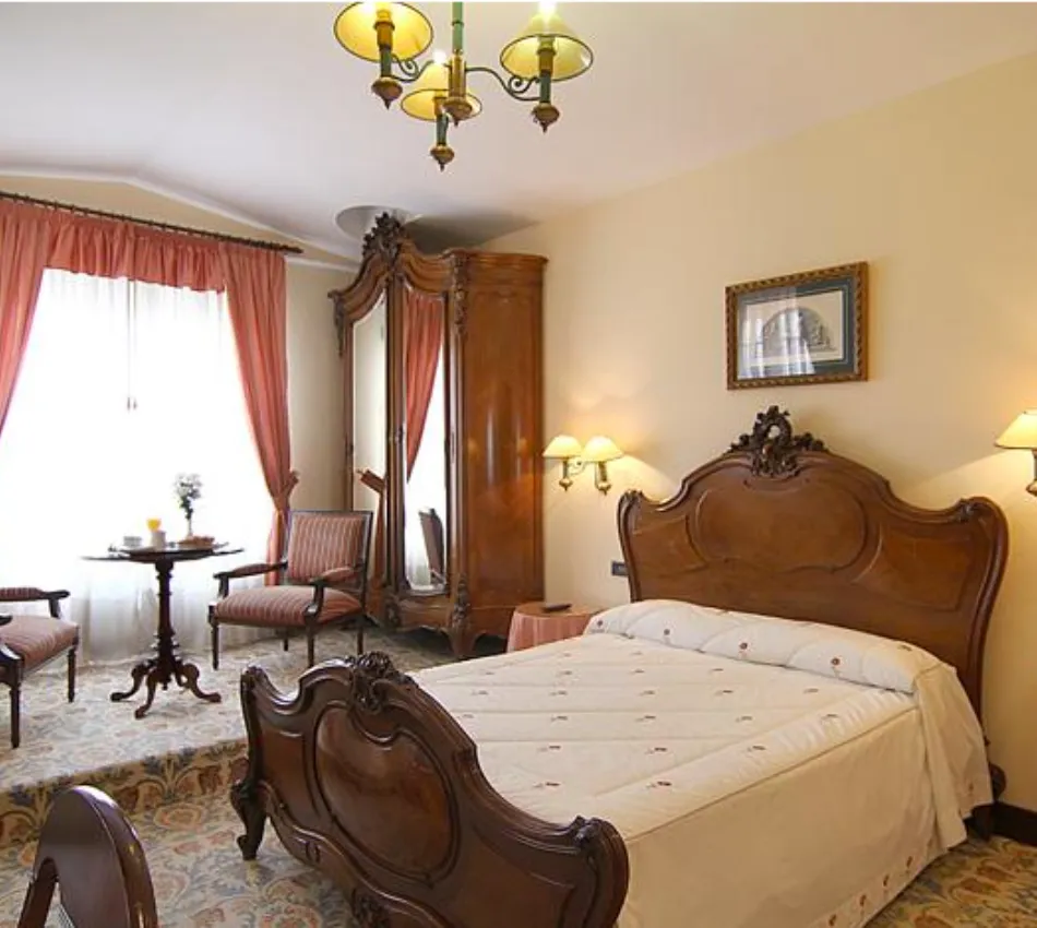 Habitaci&oacute;n con cama de madera tallada, espejo, mesa y sillas, decoraci&oacute;n cl&aacute;sica y cortinas rosadas.