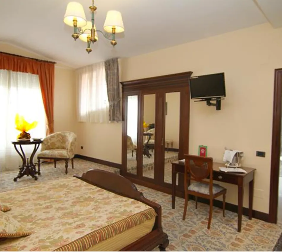 Habitaci&oacute;n cl&aacute;sica con cama, escritorio, silla, espejo y televisor en la pared.